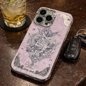 May include: A light pink phone case featuring a silver cherub design and a silver frame. The case is decorated with small pink crystals. The text "Castiel's Angels" is visible on the case. The phone case is next to a car key.
