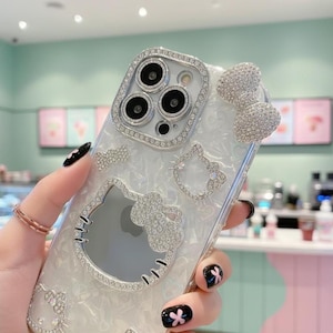 May include: Clear phone case with a silver rhinestone design featuring a bow, a Hello Kitty outline, and a rhinestone-encrusted frame around the camera. The case has a pearlescent background and is held by a hand with black nail polish.