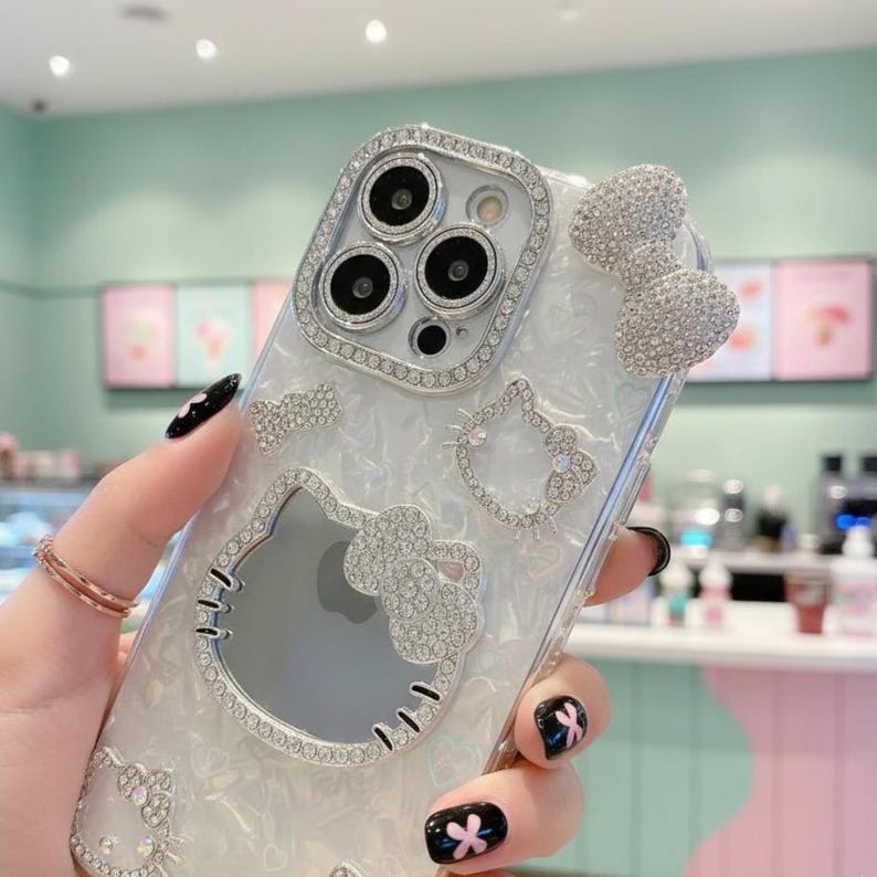 May include: Clear phone case with a silver rhinestone design featuring a bow, a Hello Kitty outline, and a rhinestone-encrusted frame around the camera. The case has a pearlescent background and is held by a hand with black nail polish.