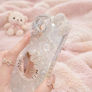 May include: A clear phone case adorned with sparkling rhinestones. The case features a bow, a cat face, and other decorative elements. The camera lens area is also embellished with rhinestones. The case is held by a person with a ring on their finger.