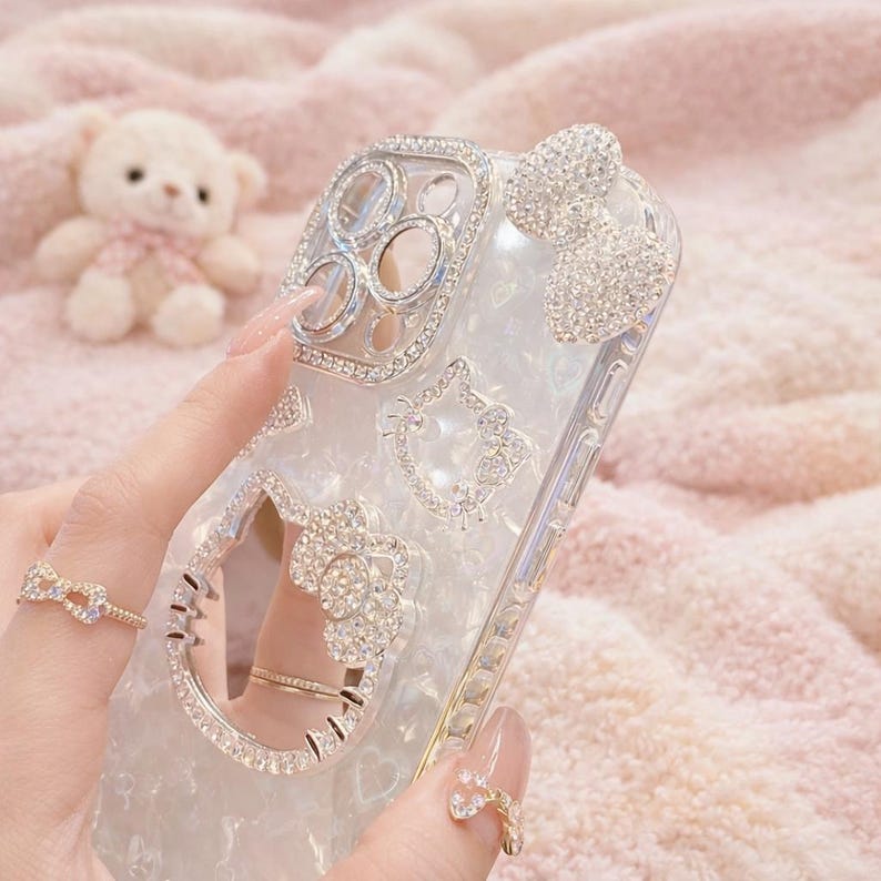 May include: A clear phone case adorned with sparkling rhinestones. The case features a bow, a cat face, and other decorative elements. The camera lens area is also embellished with rhinestones. The case is held by a person with a ring on their finger.