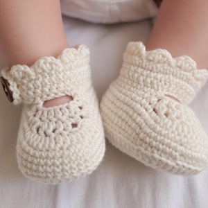 Puede incluir: Un par de patucos de bebé de ganchillo color crema. Cada patuco presenta un borde festoneado, un pequeño cierre de botón y un recorte decorativo en la parte superior. Los patucos están hechos de hilo suave y texturizado.