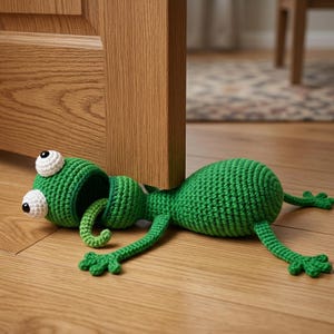 May include: A green crocheted doorstop shaped like a cartoon frog. The frog has large white and black eyes, a long tongue, and outstretched arms and legs. The doorstop is lying on a wooden floor, holding a wooden door open.