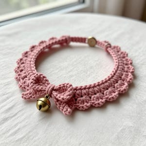 May include: A dusty pink crocheted collar featuring a scalloped edge, a small decorative bow at the front, and a round brass-toned metal bell. The collar includes a circular metal fastener at the back to secure the ends together.