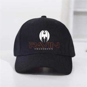May include: Black baseball cap with the word "RAVIN" in orange and white text. The cap features a white graphic of a stylized bird or bat. The text "CROSSBOWS" is below the word "RAVIN".