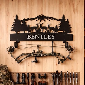 May include: A hunting-themed room with a wooden wall and shelves. A black metal sign with the name "Bentley" and a deer silhouette hangs above a bow and arrow. Camouflage backpacks, bags, and hunting gear are displayed throughout the room.