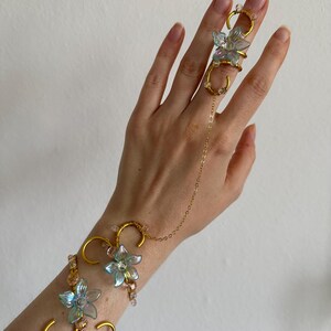 Könnte beinhalten: Eine Handkette aus goldfarbenem Metall mit irisierenden blauen blumenförmigen Anhängern und kleinen klaren Perlen. Das Schmuckstück verbindet ein Armband mit einem Fingerring durch eine Kette, ergänzt durch geschwungene Drahtelemente und Blüten.