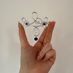 Pode incluir: Um acessório de cabelo em fio metálico prateado com um design de espiral central, dois detalhes em forma de flor branca com centros azul-escuros e uma única conta azul-escura pendente. O fio metálico forma laços e curvas simétricas.