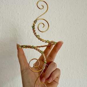 Könnte beinhalten: Ein goldfarbener Armreif aus Draht mit einem geschwungenen, schlangenförmigen Design. Der Metalldraht ist entlang seiner Länge mit kleinen, hellgrünen Perlen umwickelt und bildet zwei markante Spiralenden, die in entgegengesetzte Richtungen verlaufen.