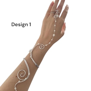 Könnte beinhalten: Ein silberfarbenes Handkettchen- und Armreif-Set mit der Aufschrift Design 1, bestehend aus drahtgewickelten Spiralen und kleinen weißen Perlen. Der Schmuck verbindet einen Ring am Mittelfinger mit einer zarten, perlenbesetzten Kette am Arm.