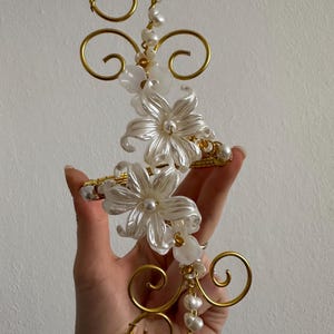 Op de afbeelding: Een decoratief haaraccessoire met goudkleurige draadkrullen en witte bloemaccenten. Het ontwerp bevat imitatieparels. Het item wordt in een hand gehouden, wat de delicate details laat zien.