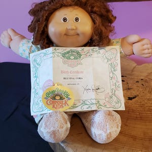 Puede incluir: Muñeca Cabbage Patch Kids con cabello rizado castaño rojizo, vestida con un vestido con estampado floral. La muñeca sostiene un certificado de nacimiento que dice "BELVINA CORA" y "SEPTEMBER 1ST". También es visible una etiqueta dorada con el logotipo de Cabbage Patch Kids.