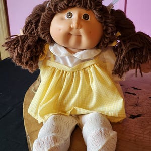 Pode incluir: Uma boneca vintage com cabelo castanho de lã em tranças, vestindo um vestido xadrez amarelo sobre uma blusa branca. A boneca tem um rosto redondo, bochechas rosadas e usa meias brancas. Um brinquedo colecionável clássico.