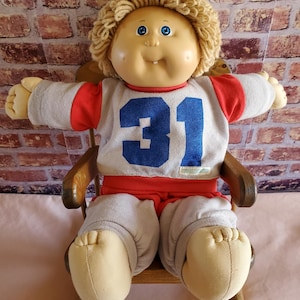 May include: A vintage Cabbage Patch Kid doll with blonde yarn hair, blue eyes, and a smiling expression. The doll is wearing a gray and red jersey with the number 31 in blue, gray pants, and is seated in a wooden chair.