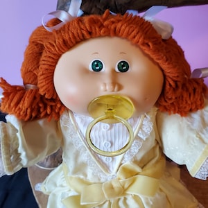 Op de afbeelding: Een vintage Cabbage Patch Kid pop met oranje garen haar, groene ogen en een gele fopspeen. De pop draagt een crèmekleurige jurk met kanten rand en een gele strik. Witte linten zijn in het haar gebonden.
