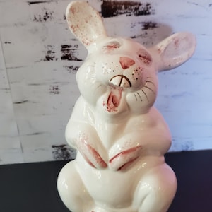 May include: A glossy white ceramic rabbit figurine with a wide, open-mouthed expression. The rabbit features hand-painted pink accents on its ears, nose, paws, and feet, with gold-colored detailing on its two front teeth and whiskers.