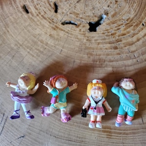 May include: Four small plastic figurines of children with round faces and yarn-like hair. The first wears a purple tutu, the second has roller skates and a teal outfit, the third is dressed as a nurse with a stethoscope, and the fourth wears teal athletic clothing.
