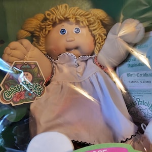 May include: A vintage Cabbage Patch Kids doll from 1985, with blonde yarn hair, blue eyes, and a white dress with black trim. The doll has a tag with the Cabbage Patch Kids logo and a birth certificate.