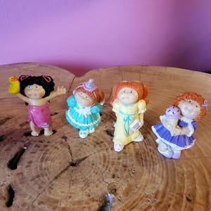 May include: Four small Cabbage Patch Kids figurines. The first has black hair and a pink towel, the second has auburn hair and a teal dress, the third has orange hair and a yellow dress, and the fourth has orange hair and a purple dress.