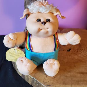 May include: A plush doll with light skin, a round face, and curly white hair. The doll wears bright blue shorts with rainbow-striped suspenders. A yellow, heart-shaped tag is attached to the doll's arm, featuring printed text about the Koosa character.