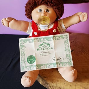 Op de afbeelding: Een Cabbage Patch Kids pop met bruin wollen haar, gekleed in een wit shirt en een rode corduroy tuinbroek, met een gele fopspeen in de mond. De pop wordt vergezeld door een geboortecertificaat voor Bo Theodore, gedateerd op 1 oktober.