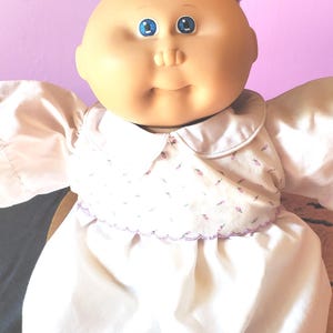 May include: A bald doll with a round, peach-toned face, blue eyes, and a small nose. The doll wears a white dress featuring a collar and a bodice decorated with a small, repeating purple floral pattern, finished with a scalloped purple hemline.