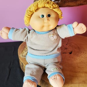 May include: A vintage Cabbage Patch Kid doll with blonde yarn hair, green eyes, and a soft, round face. It wears a grey outfit with blue trim and a small cat patch. The doll is posed with arms outstretched.