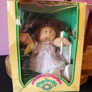 Könnte beinhalten: Eine Vintage Cabbage Patch Kids Puppe in ihrer Originalverpackung. Die Puppe hat braune Haare und ein rosa Kleid und hält eine Zahnbürste. Die Box ist gelb und grün mit den Worten "The Official Cabbage Patch Kids" und "I'm unique, I'm adoptable!"