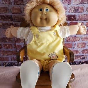 Puede incluir: Una muñeca Cabbage Patch Kid vintage con cabello rubio y esponjoso, ojos azules y una expresión sonriente. La muñeca lleva una camisa blanca y un peto amarillo con una aplicación de ratón, sentada en una silla de madera. Los zapatos de la muñeca son blancos.