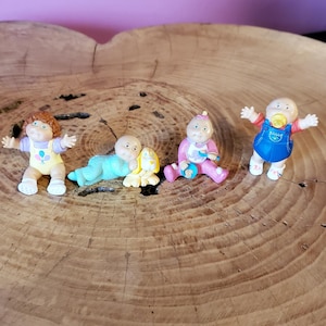 May include: Four small Cabbage Patch Kids figurines. One wears a yellow outfit with balloons, one is a baby in blue pajamas with a yellow dog, one is in a pink outfit with a bib, and one is in blue overalls with a yellow pacifier in its mouth.