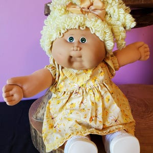 May include: A doll with light skin, green eyes, and curly pale yellow yarn hair adorned with a tan ribbon bow. The doll wears a yellow dress featuring a pattern of small orange flowers and dragonflies, paired with white socks and white shoes.