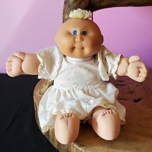 Könnte beinhalten: Eine Stoffpuppe mit einem runden, gebräunten Gesicht, leuchtend blauen Augen und einem kleinen Büschel aus blondem Garn auf dem Kopf. Die Puppe trägt ein weißes Kleid mit kurzen, gerüschten Ärmeln, die mit Lochstickerei verziert sind.