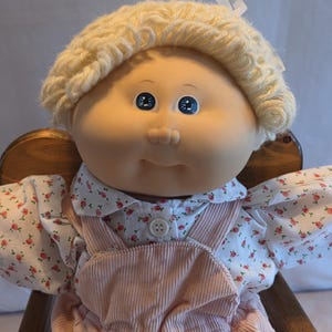 Peut inclure: Une poupée vintage avec un visage en vinyle doux, des yeux bleus et des cheveux blonds en laine. La poupée porte un chemisier à imprimé floral et une salopette rose en velours côtelé. Un ruban blanc est noué dans les cheveux. La poupée est assise dans une chaise en bois.