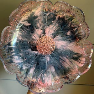 Könnte beinhalten: Ein blumenförmiges Harztablett mit einem wirbelnden Muster aus schwarzen und zartrosa Pigmenten. Die Mitte ist mit dichtem roségoldenem Glitzer gefüllt, während die gewellten Ränder mit verstreuten roségoldenen Flocken im klaren Harz verziert sind.