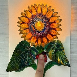 Op de afbeelding: Een handgeschilderd zonnebloemkunstwerk met een gloeiend licht. De zonnebloem heeft oranje bloemblaadjes, een blauw en geel centrum en groene bladeren met een lieveheersbeestje. Het kunstwerk staat op een witte achtergrond.