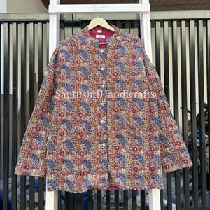 Peut inclure: Veste matelassée à motifs floraux rouges, bleus et beiges. La veste a un col mandarin et est boutonnée. Le texte "Santushti Handicrafts" est visible sur le devant.