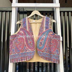 May include: A sleeveless vest with a paisley pattern in shades of red, blue, and purple. The vest has a tan-coloured interior and is hanging on a wooden hanger. The text "Santushti Handicrafts" is visible.