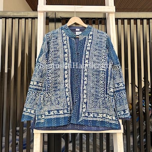 May include: A blue jacket with a white patterned design. The jacket has a button-up front and long sleeves. The pattern includes paisley and floral elements. The jacket is hanging on a wooden hanger against a white wooden structure.