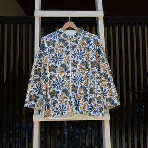 May include: A long-sleeved shirt with a floral design in blue, gold, and white. The shirt is buttoned up and hangs on a wooden hanger. The background includes a white wooden frame and a black metal fence.