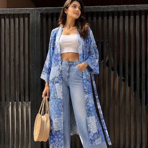 Op de afbeelding: Een lange blauwe kimono met een wit paisley patchworkpatroon, gedragen over een wit kort hemdje en een lichtblauwe wijde spijkerbroek. De outfit wordt gecombineerd met witte teenslippers en een ronde geweven strooien handtas met twee hengsels.
