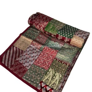 Puede incluir: Un edredón de retazos con una variedad de telas estampadas en tonos de rojo, verde y dorado. El edredón es rectangular con una sección enrollada en la parte superior. Los bordes son de color burdeos oscuro.