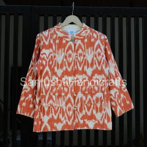 May include: A jacket with an orange and white ikat pattern, long sleeves, and two front pockets. It features a button-up front and a mandarin collar. The text "SantUshtivhandicrafts" is printed on the front.