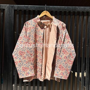 May include: A beige, open-front jacket with a floral pattern in red and blue. The jacket is hanging on a wooden hanger against a dark metal gate. The interior lining is a solid peach colour.