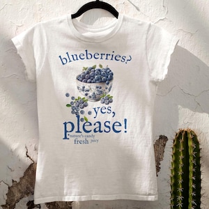 May include: White t-shirt with a blue graphic of blueberries in a decorative bowl and the words "blueberries? yes, please!" The text "nature's candy fresh juicy" is also printed on the shirt. A cactus is in the background.