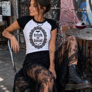以下が含まれることがあります： 白と黒のラグランTシャツ。中央には黒い装飾的な楕円形のフレームがあり、その中に「trick or treat」という文字と小さなコウモリのシルエットが描かれています。Tシャツは短い黒い袖と、丸いネックラインの白い身頃が特徴です。