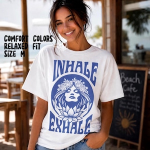 Pode incluir: Uma t-shirt branca de manga curta com uma impressão gráfica azul apresentando um rosto estilizado rodeado por flores e pétalas de lótus. O texto "INHALE" está impresso acima do gráfico e "EXHALE" abaixo. A t-shirt tem a etiqueta Comfort Colors.
