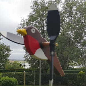 May include: A wooden bird garden spinner featuring a brown body, a bright red chest, a white belly, and a yellow beak. The bird has a black and white wing and a black propeller attached to its side, mounted on a thin metal rod for outdoor display.