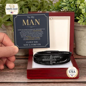 Peut inclure: Un bracelet en cuir noir à plusieurs brins avec une croix en métal au centre, présenté dans une boîte en bois sombre verni. Une carte bleu marine porte le texte : To My MAN, WHEN YOU WEAR THIS BRACELET, REMEMBER THIS LIFE MAY HOLD MANY UNCERTAINTIES, BUT MY LOVE FOR YOU IS NOT ONE OF THEM. THIS BRACELET REPRESENTS THE STRENGTH WE SHARE, THE FUTURE WE'RE BUILDING, AND THE PROMISE I CHOOSE EVERY DAY. YOU ARE CHERISHED, CHOSEN, AND IRREPLACEABLE IN MY LIFE. I LOVE YOU NOW & FOREVER.