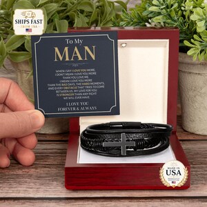 May include: A black leather multi-strand bracelet featuring a central metal cross charm, presented inside a polished dark wood box. A navy blue card is included with the text To My Man and a printed message about love and commitment.