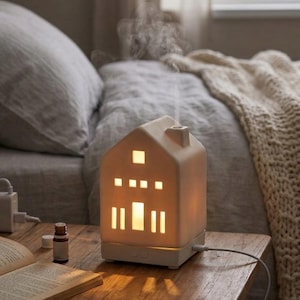 Può includere: Un diffusore di oli essenziali in ceramica bianca a forma di casa che emette un sottile getto di vapore dal camino. La struttura presenta finestre e una porta ritagliate che si illuminano con una luce calda interna, alimentato da un cavo bianco.